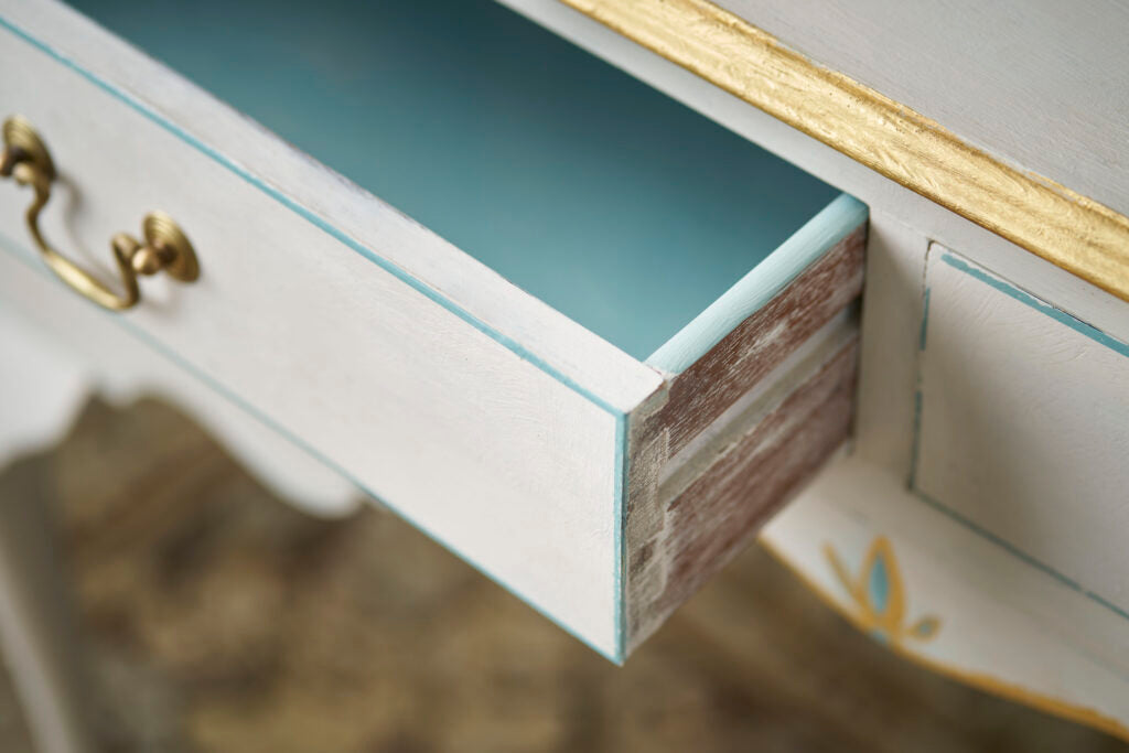 Close-up of a wooden drawer painted with Annie Sloan Chalk Paint in Rococo White with a gold handle and decorative trim.